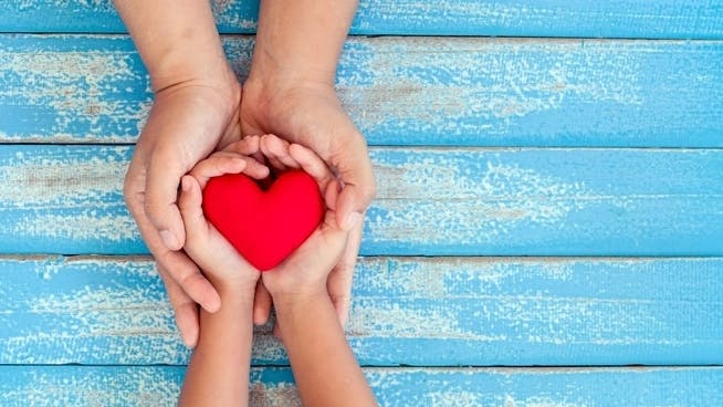 Photo from above a child's hands inside a parent's hands holding the shape of a red heart. Background is a blue wooden table.