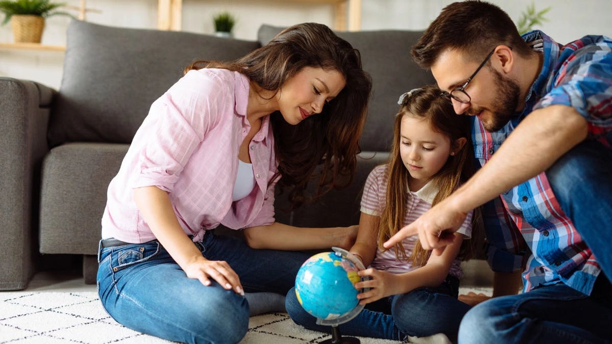 Two parents and child sitting on a carpet in front of a couch pointing at a small globe 