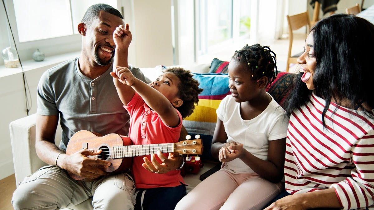 A father sits on a couch playing a small guitar with his son, daughter, and wife. 